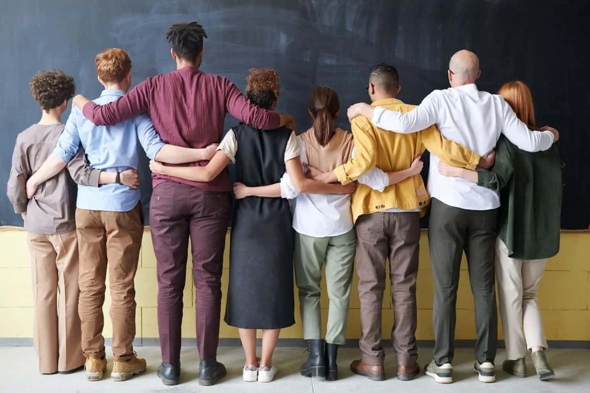 Diverse group of friends standing in a line with arms around each other facing a chalkboard, representing the close-knit friendships found in sitcoms like New Girl