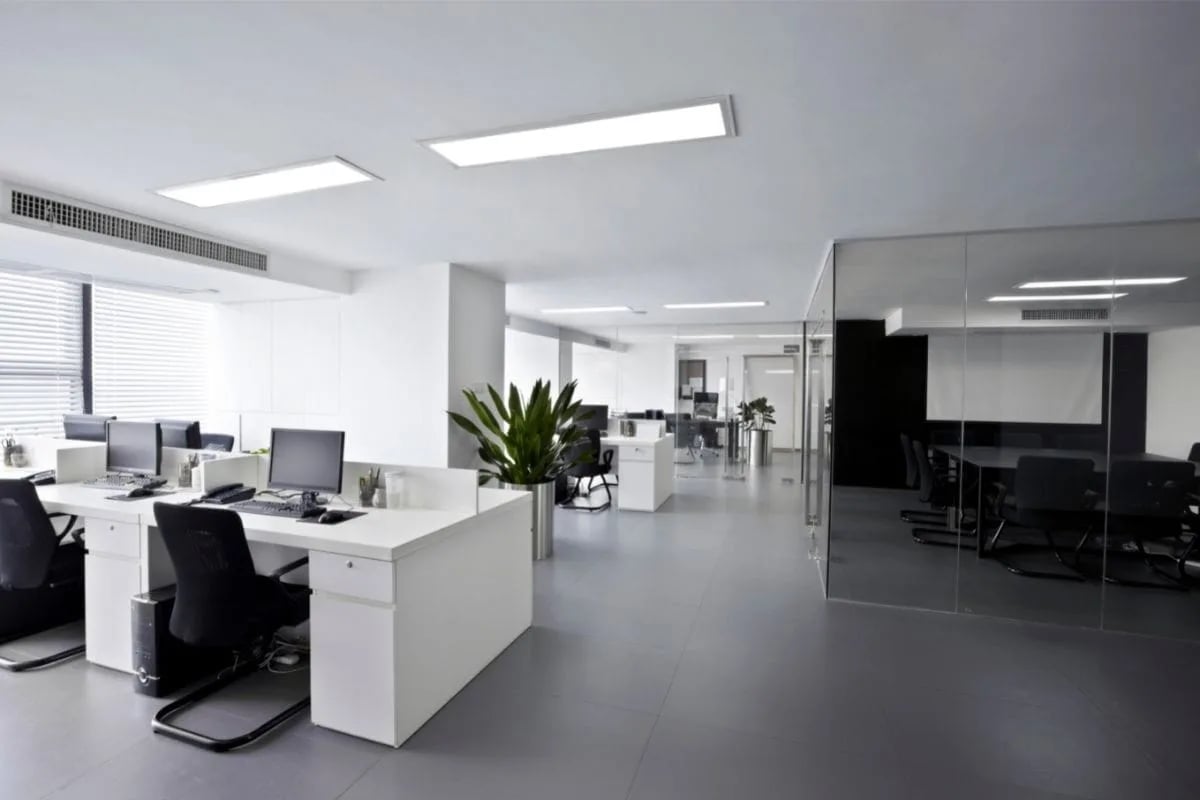 Modern open-plan office workspace with white desks, black chairs, computer monitors, and a glass-walled conference room reminiscent of settings from TV shows like The Office