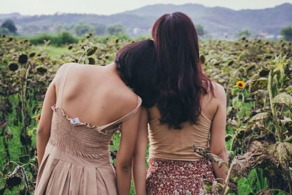 Two women with dark hair embracing in a sunflower field, scenic mountain background, peaceful outdoor setting.