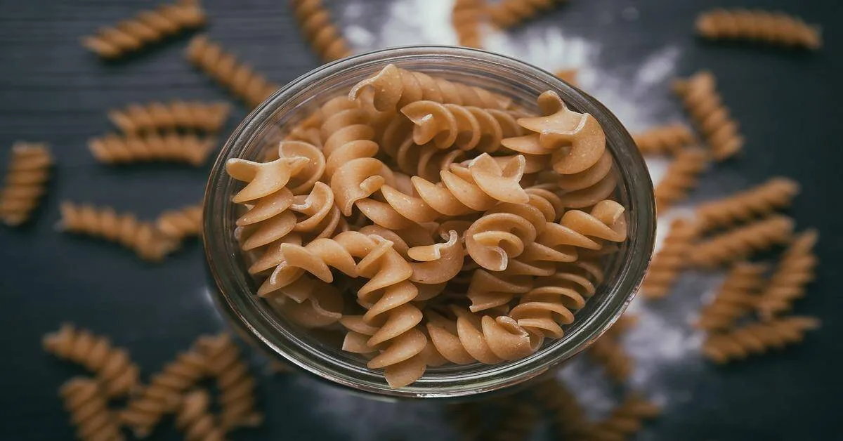 Close-up of fusilli pasta in a glass jar with some scattered on a dark surface.