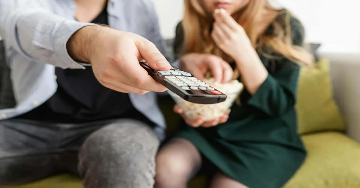 Close-up of people watching TV together, holding a remote and snacking.
