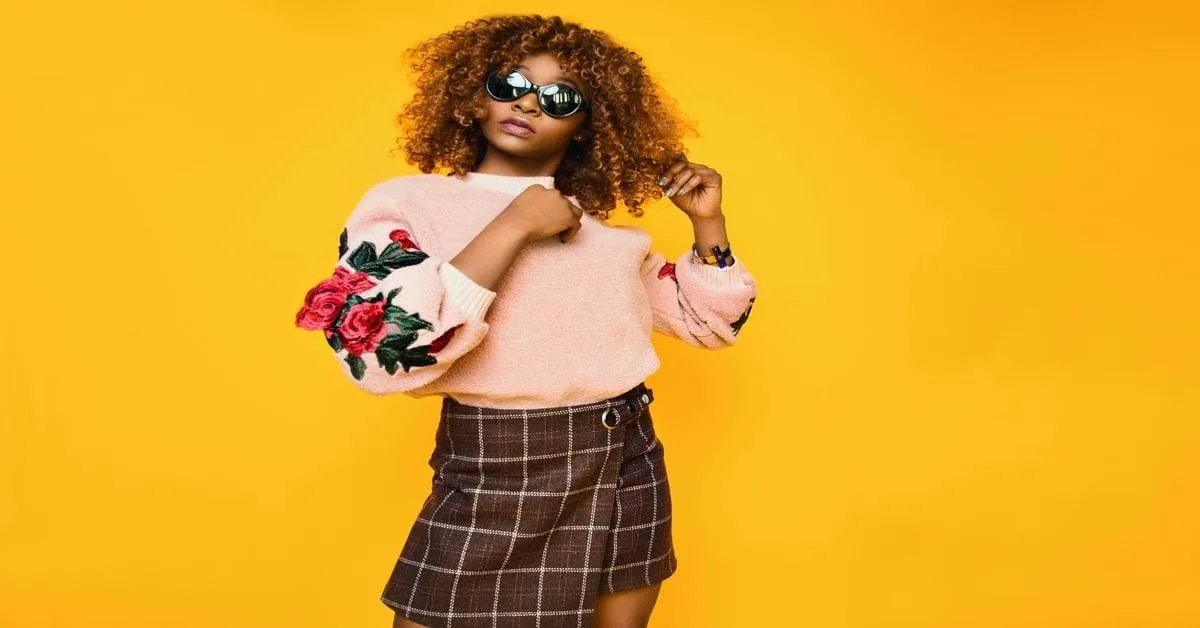 Stylish young woman in trendy sweater, checkered skirt, and sunglasses poses against yellow background.