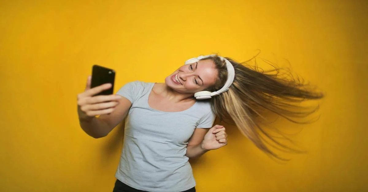 Woman enjoying music with wireless headphones, taking a selfie and dancing on bright yellow backdrop.