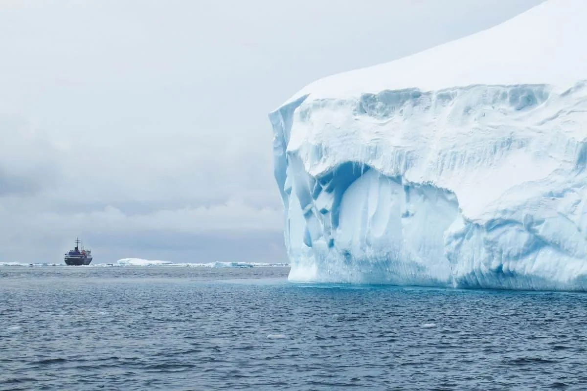 Massive arctic iceberg towering over dark ocean waters with a distant expedition ship navigating through icy polar seas under an overcast sky