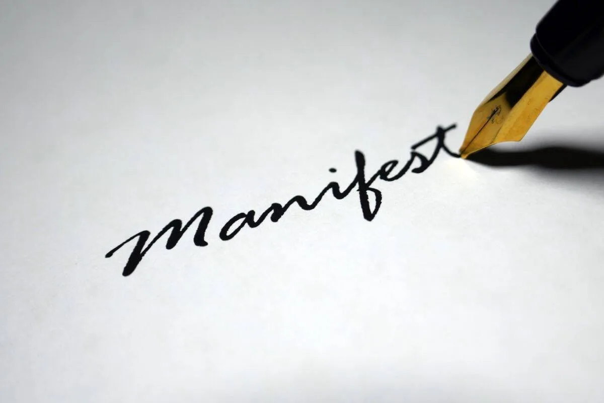 The word Manifest written in elegant cursive calligraphy with a gold-tipped fountain pen on white paper