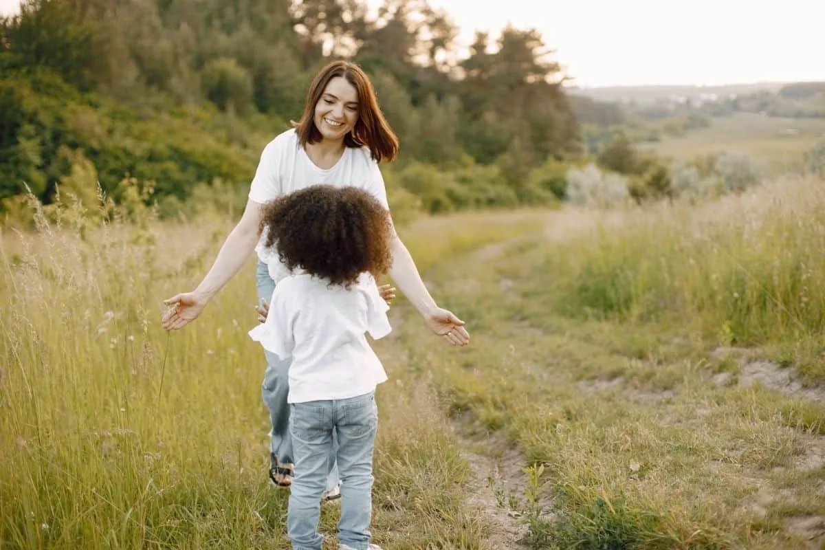 Smiling mother and young daughter with curly hair embracing on a grassy countryside path at golden hour, evoking the mother-daughter bond central to shows like Ginny and Georgia