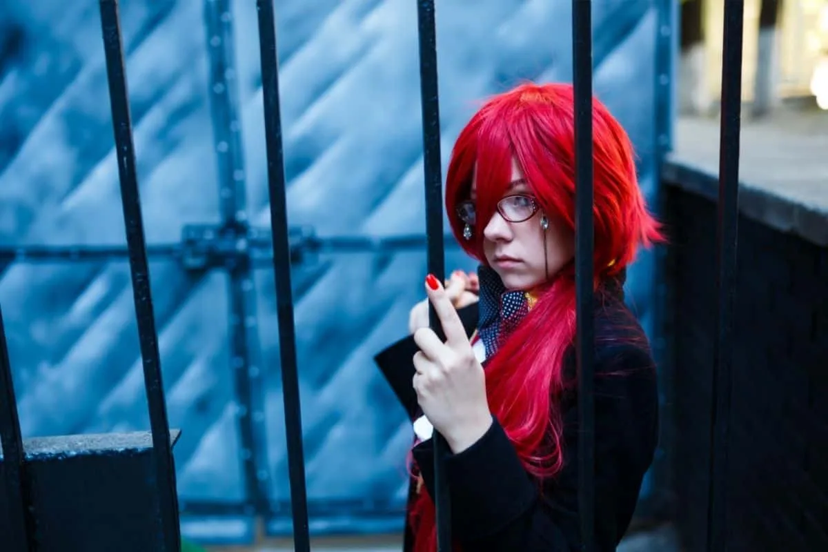 Anime cosplayer with bright red wig and glasses posing behind iron bars in a My Hero Academia inspired costume for shows like Invincible