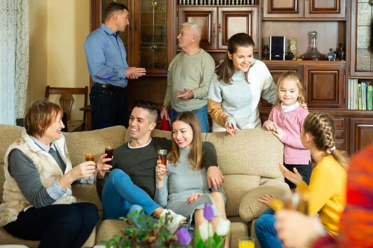 Multi-generational family gathering in a cozy living room with grandparents, parents, and children laughing and enjoying drinks on a sofa, reminiscent of shows like Modern Family