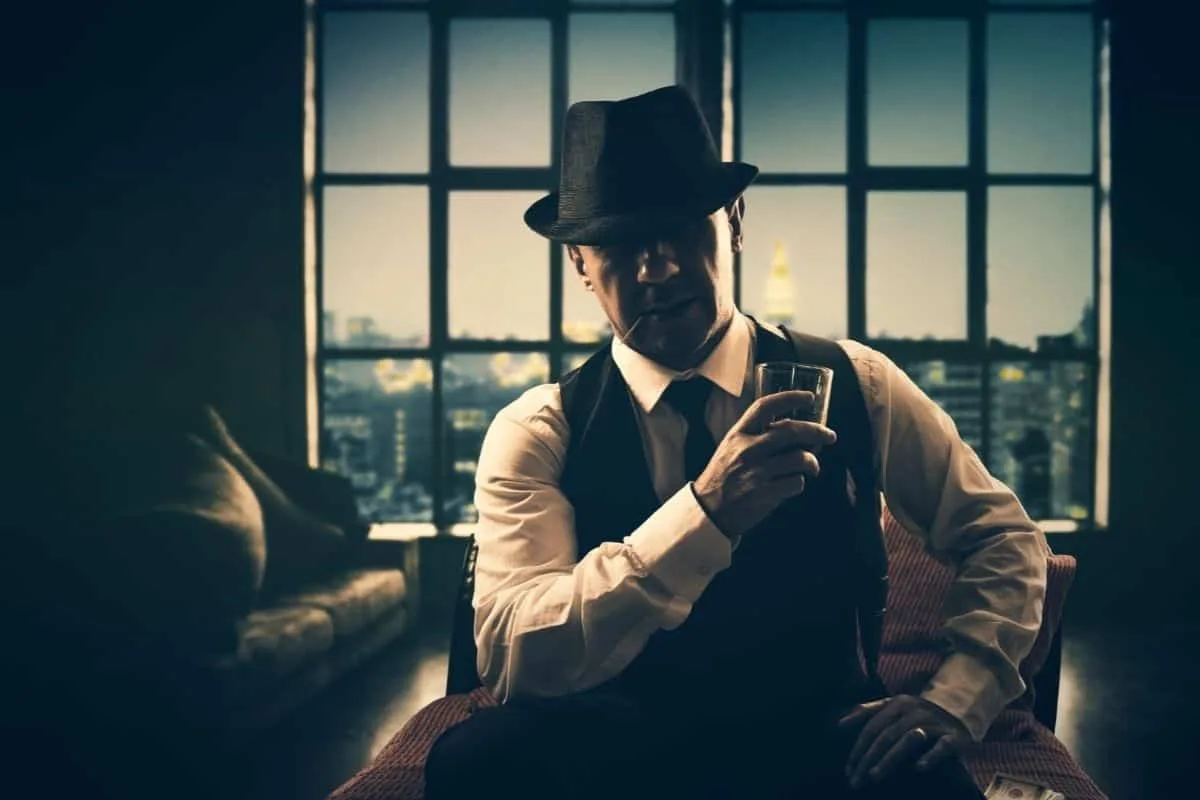Mysterious man in fedora hat and vest holding a whiskey glass in a dimly lit room with city skyline at night representing crime drama shows like Power
