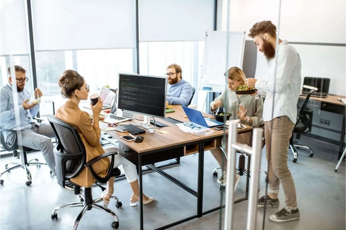 Group of coworkers chatting and eating lunch together at their desks in a modern open-plan office reminiscent of The Office TV show workplace comedy setting